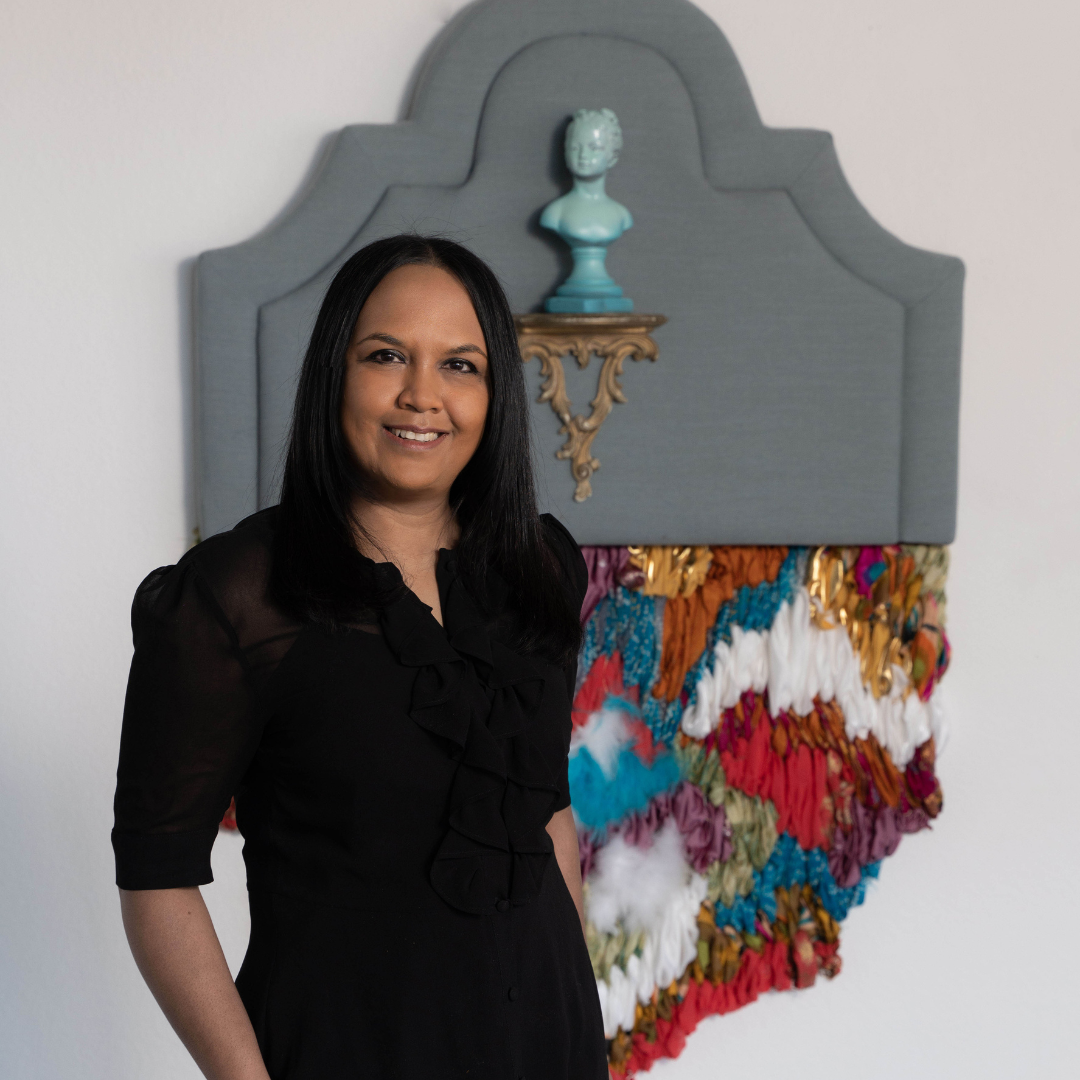 Portrait of artist Suchitra Mattai sporting a black shirt and smiling softly. Suchitra is standing next to a mixed media artwork.