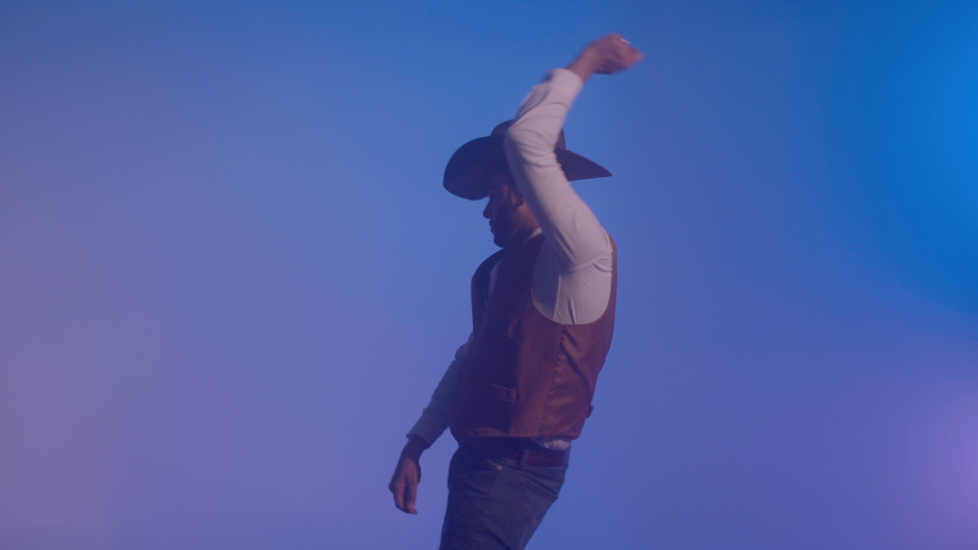 Still from a video of a cowboy against a bluish, purplish sky. The cowboy is sporting a cowboy hat and vest, and has one arm up in the air. 