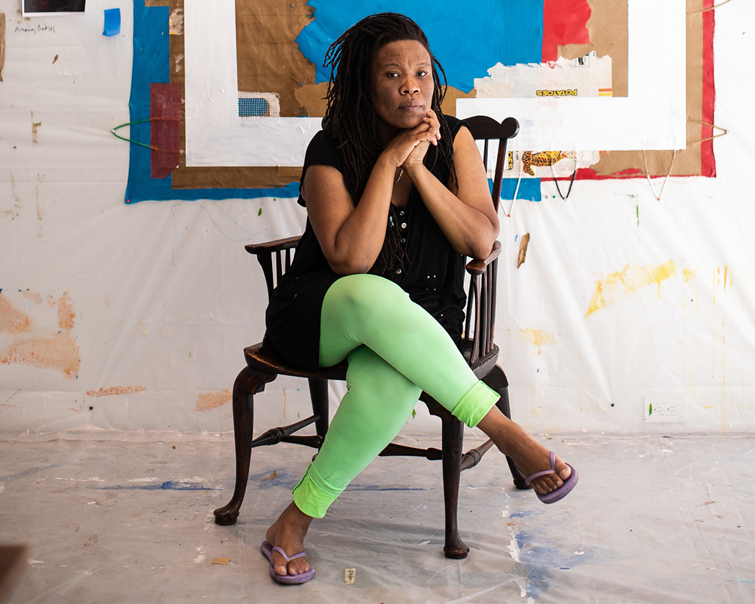 Artist Tomashi Jackson sitting in a chair with her legs crossed. She is sitting in what looks like a colorful studio.