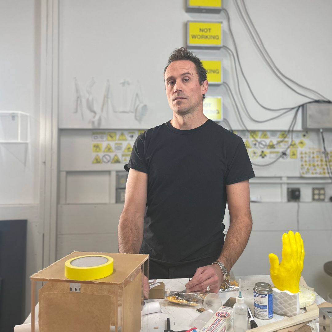 Portrait of artist Trey Duvall in his studio. He’s surrounded by various materials, included a yellow sculpture of a hand.
