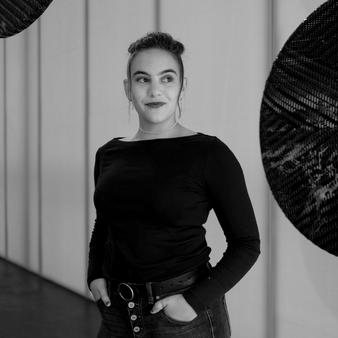 Black and white portrait of person named Zaida Sever, standing next to a sculpture where large planet-like shapes suspend from the ceiling.