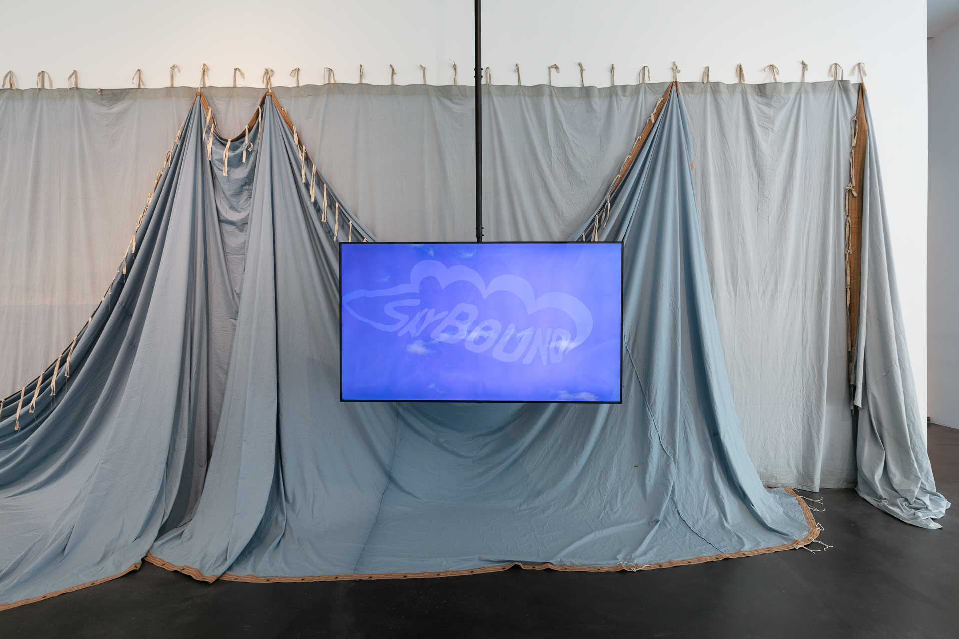 Television suspended from a ceiling in a gallery. Behind the TV is a large blue theater curtain.