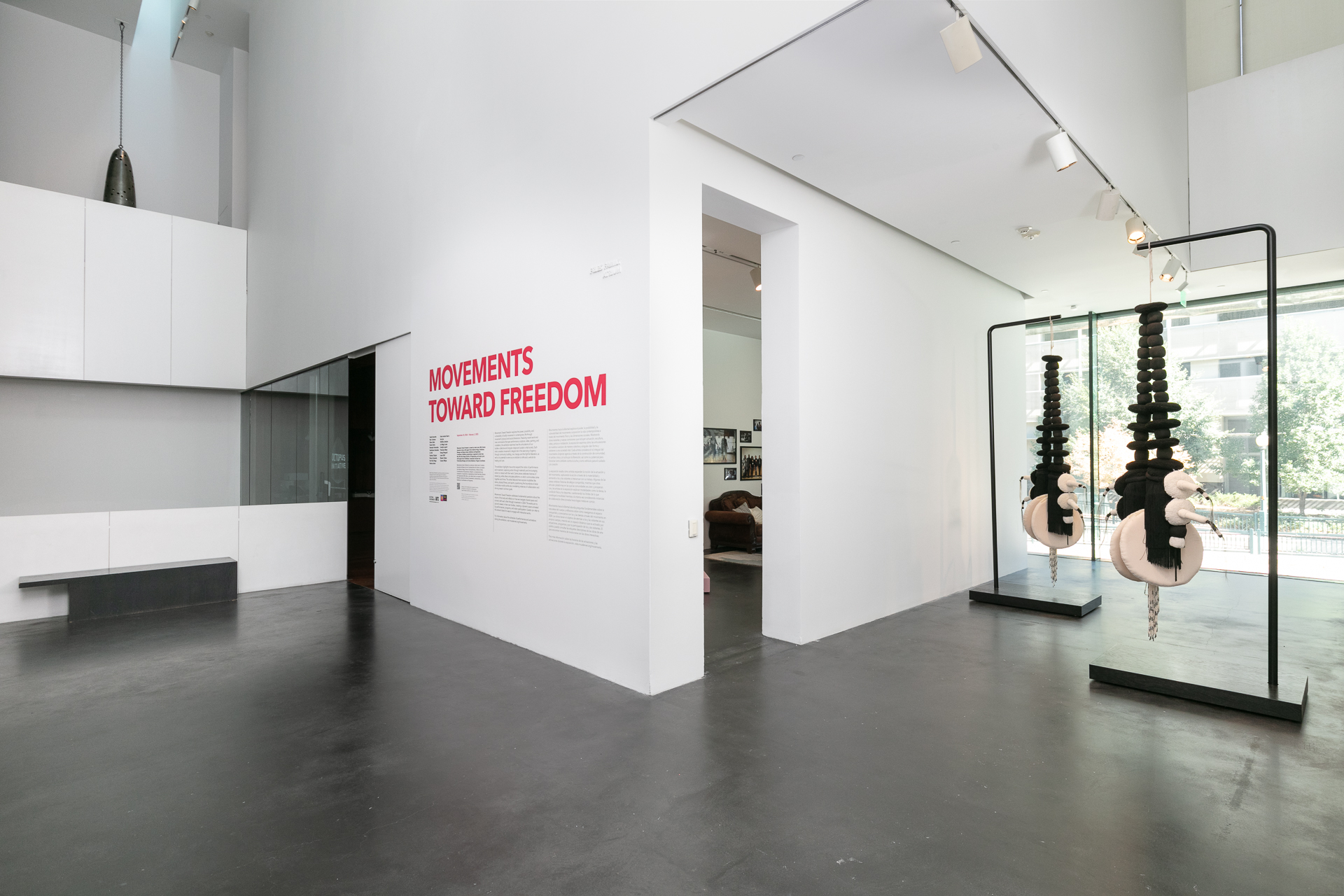 Large atrium in a museum. In the image is wall text describing the exhibition, a soft sculpture in a gallery, and a view of a bell hanging from the second floor. 