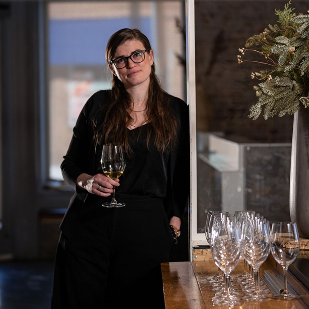 Photo of Caroline Clark holding a wine glass, wearing a black outfit, and leaning up against a restaurant wall. 