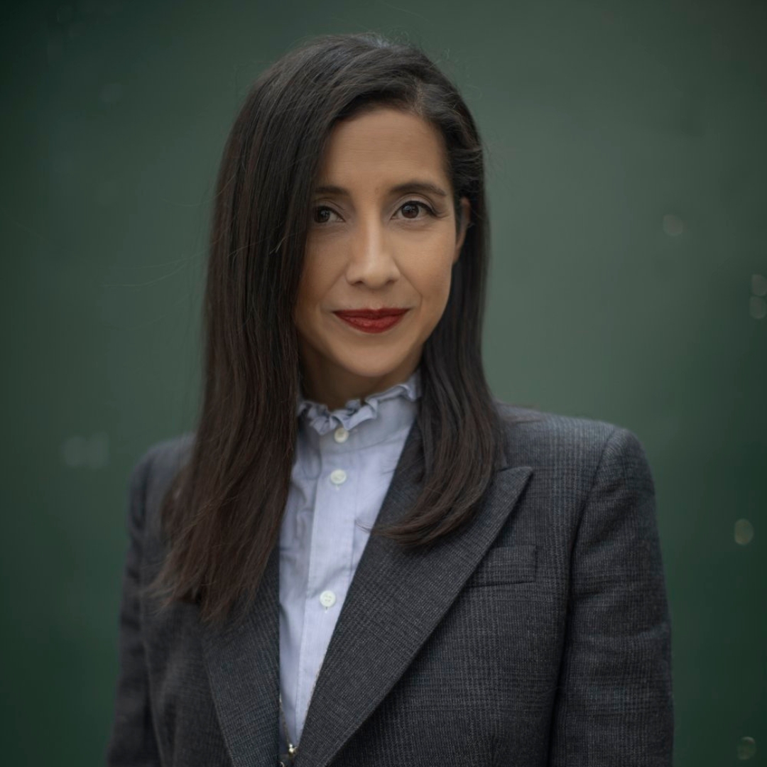 Karla Martínez de Salas sporting dark straight hair, a blazer, and standing in front of a forest green background.