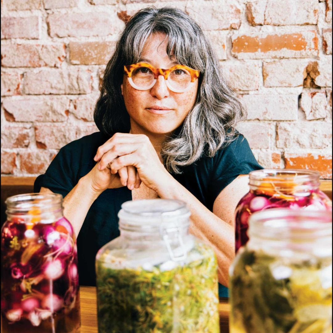 Portrait of Mara King sporting big glasses and sitting in front of jars of fermented foods.