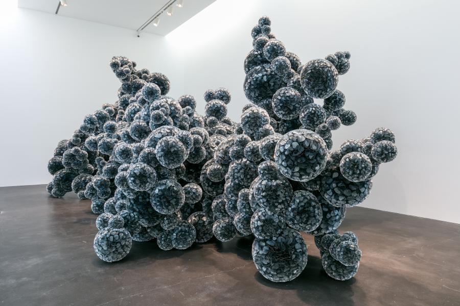 A large sculpture rests in a gallery space. It is composed of mylar spheres resulting in a large black amorphous shape. 