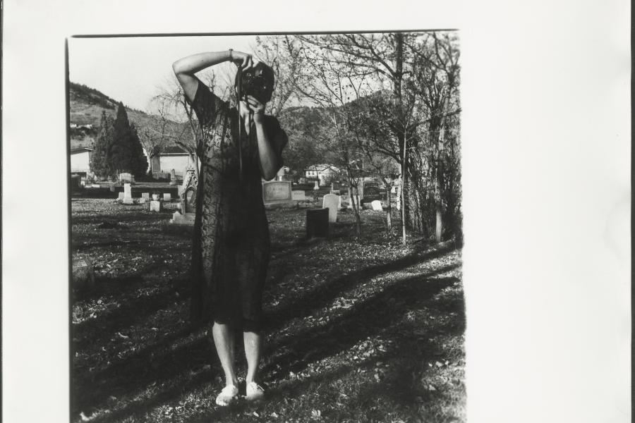 Francesca Woodman, Untitled photograph, circa 1975-1978. Gelatin silver print. A woman is standing in a graveyard in a sheer black dress. She is holding a camera up to her face at an angle. 