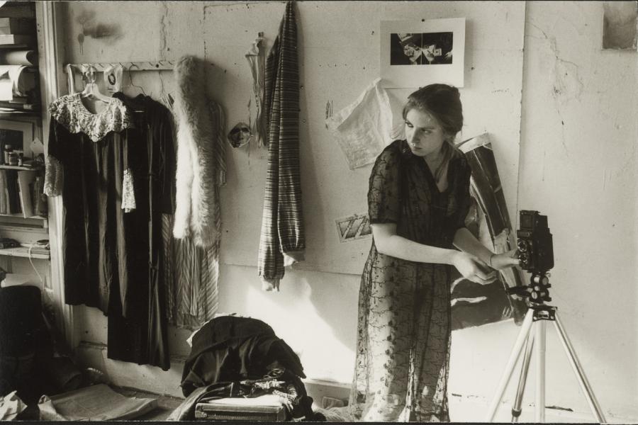 George Lange, Untitled photograph, circa 1975-1978. Gelatin silver print. Candid shot in a sepia tone of a woman setting up a camera on a tripod. She is indoors and wearing a sheer dress. Behind her is a wall where clothes and photographs hand. The floor is cluttered with various belongings