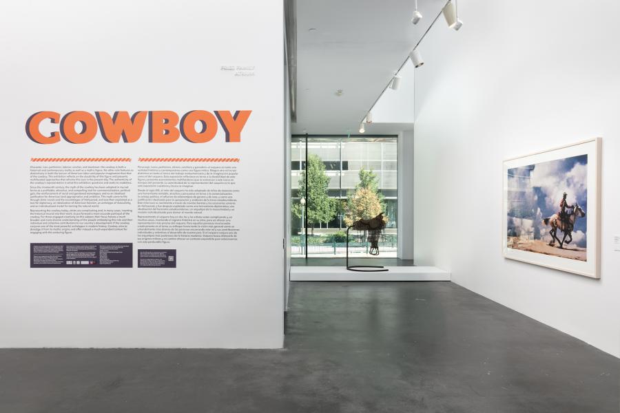 Gallery featuring wall text that reads, "Cowboy" in orange font. There is an artwork that looks like a saddle and a photograph of a cowboy riding a horse in the gallery.