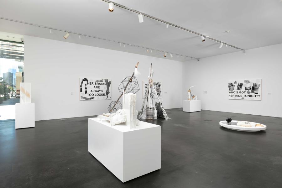 Gallery at MCA Denver with white walls and concrete floors. The gallery has works on the ground and hung on the wall. The works on the wall look like collages and there is a teepee-like structure and pedestals with monochromatic sculptures on the floor.