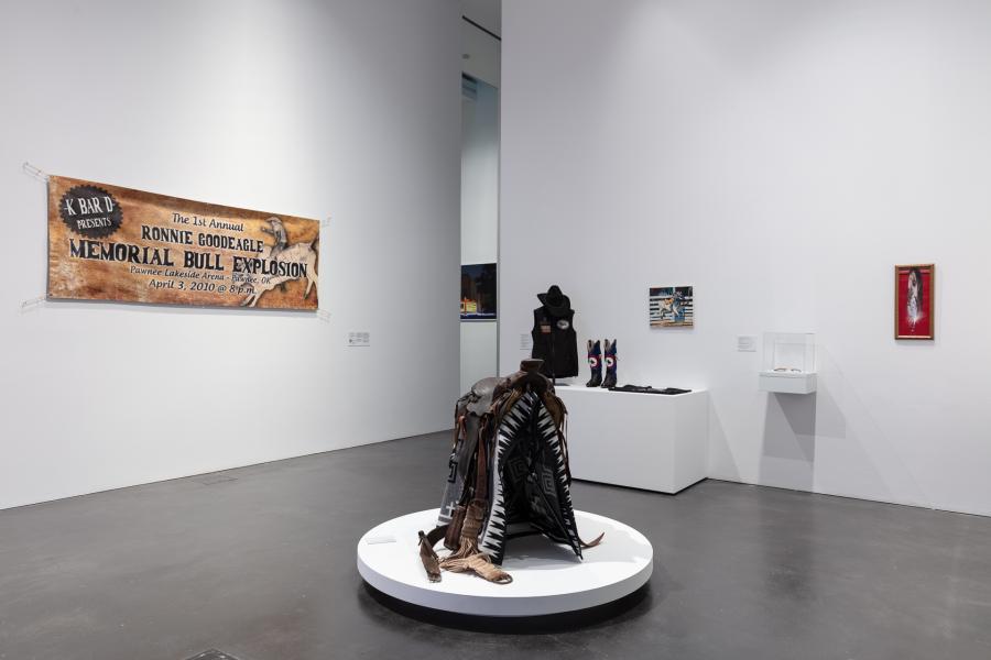 Gallery featuring a saddle in the center of the space, surrounded by cowboy objects such as cowboy boots and a cowboy hat.