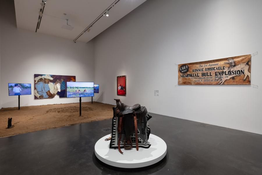 Gallery featuring a saddle on display in the center of the room. Behind the saddle is an installation featuring dirt on the ground, televisions mounted in the dirt, and artwork on the walls.