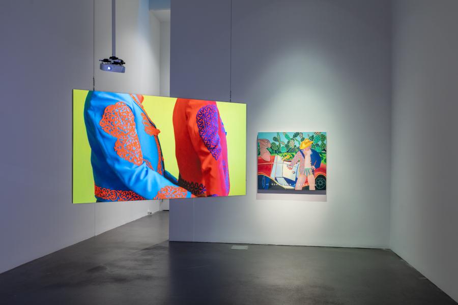 Gallery featuring a video on a screen that is suspended from the ceiling. There is a colorful painting behind the screen that is hung on the wall. 