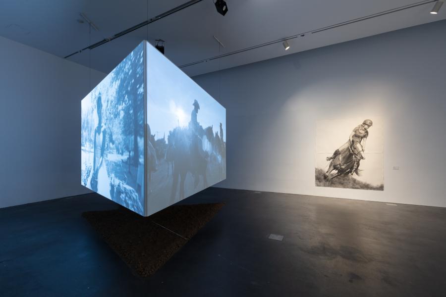 Gallery featuring a large, three-channel video suspended from the ceiling. In the gallery there is also a large drawing of a girl riding a horse.