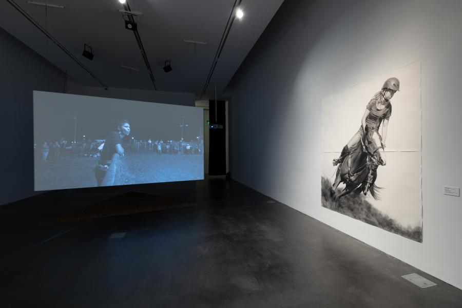 Gallery space featuring a large video suspended from the ceiling. In view is a large drawing of a girl on a horse.
