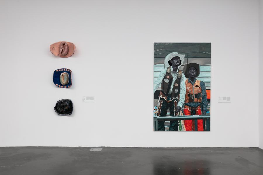 Gallery featuring three ceramic cowboy hats hung on a white wall and a large painting of two Black cowboys.