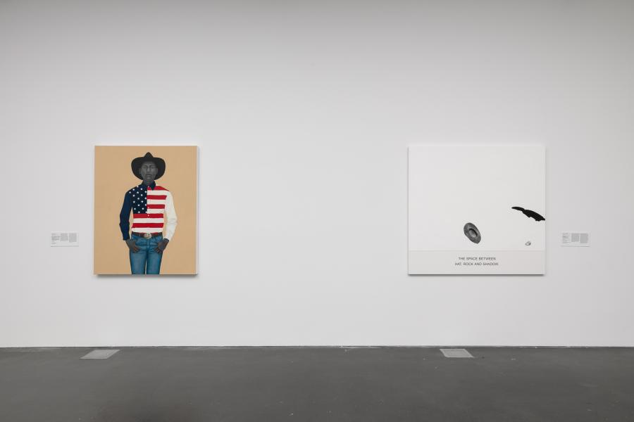 Two paintings hung on a white wall in a gallery. One painting features a Black cowboy wearing an American flag shirt. The other is white and black and minimalistic. 