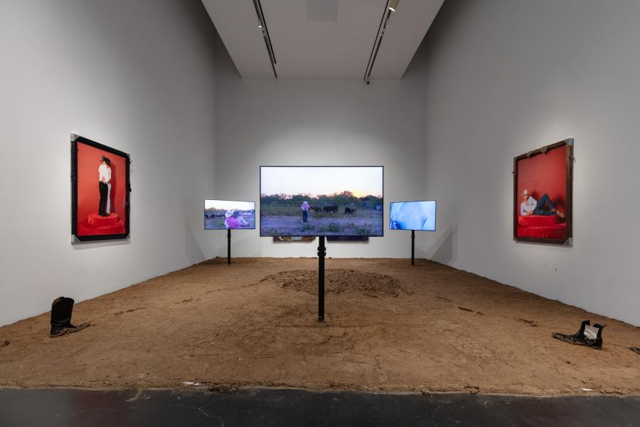 Installation featuring dirt on the group, three televisions mounted in the dirt, and artwork of gay cowboys on the walls.