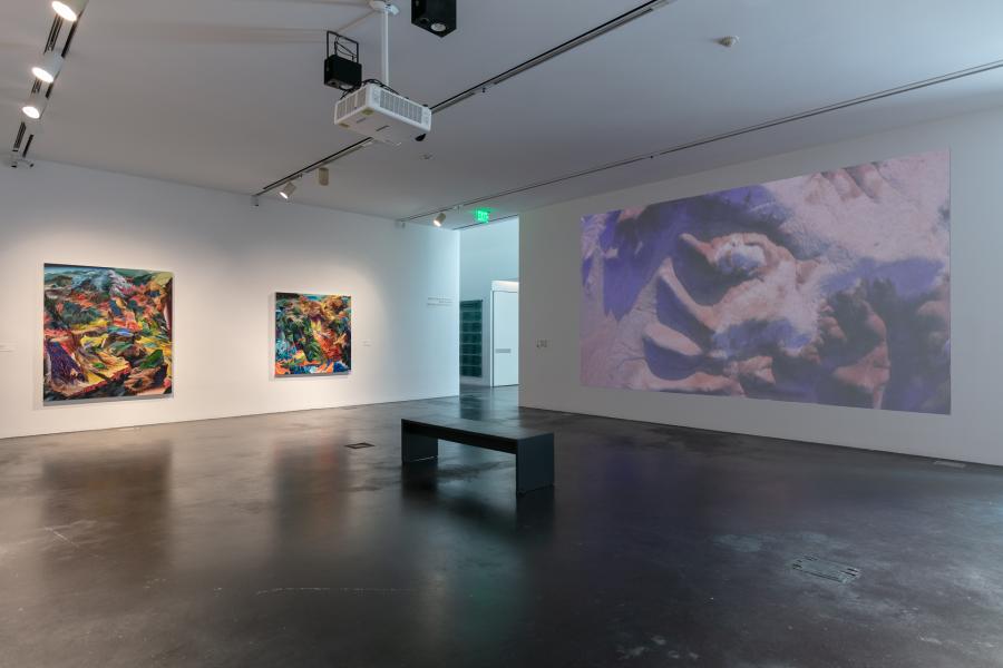 Gallery featuring two large colorful paintings and a video projection featuring a bird's eye view of landscape scenes. 