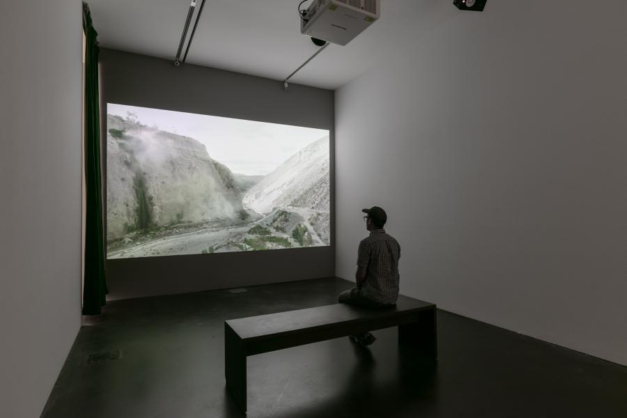Video installation in a dark gallery. There is a person watching the video on a bench.