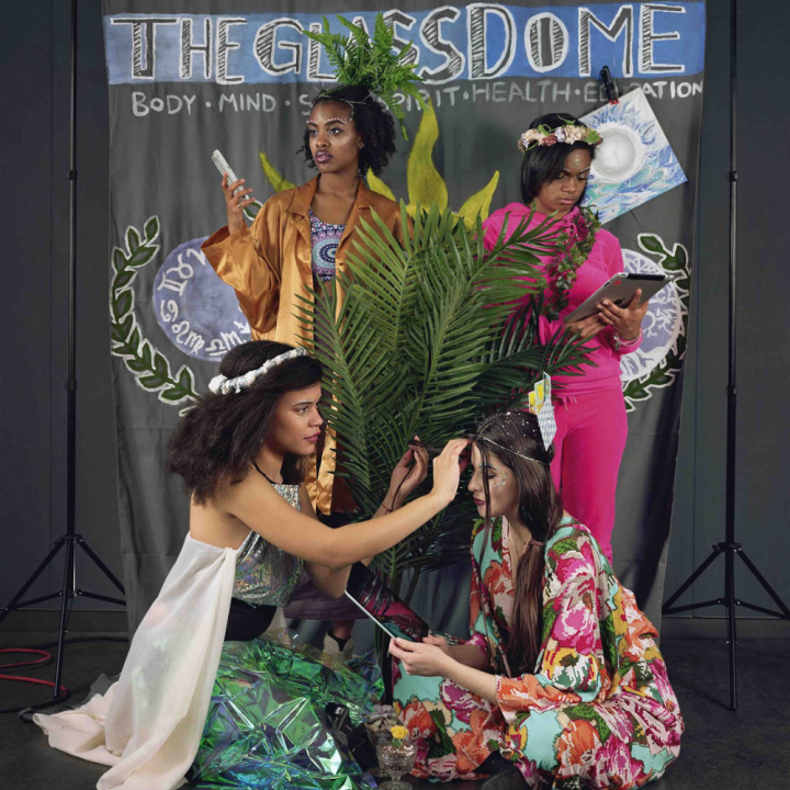 Four young women of different racial backgrounds surround a fern in an indoor space. They are dressed colorfully while donning avante garde makeup. They are posed in front of a hand painted background with imagery of plants and zodiac symbols while two of them look at iPads.