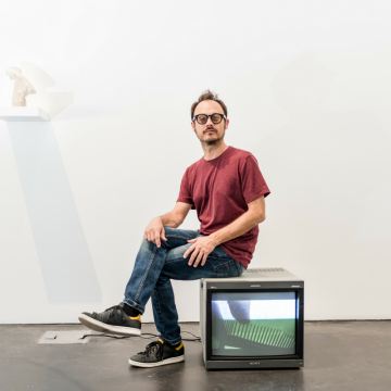 A shot of a man sitting on an old boxy tv in a gallery space. 