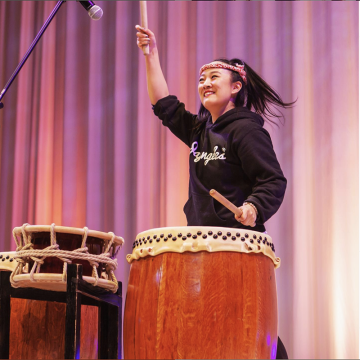 Person standing in front of pink curtains, smiling big and beating a drum.