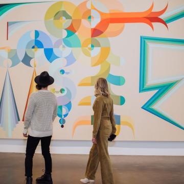 Two people with their backs towards the camera, looking at a massive, colorful, abstract artwork.