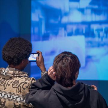 Two people standing with their backs facing the camera, photographing a blue projector on one of their phones. 