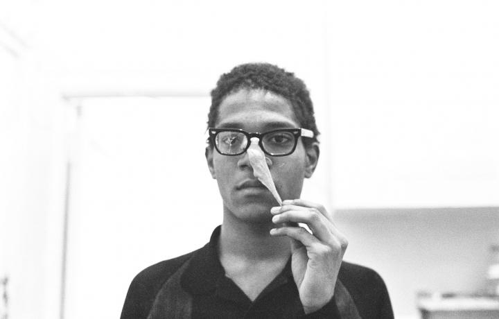 A grainy black and white portrait of Jean-Michel Basquiat in an indoor space. He is wearing a black button up shirt, black glasses with one lens fractured. His hair is short and tight. He is pulling a clay-like substance off his face while looking neutrally into the camera.  