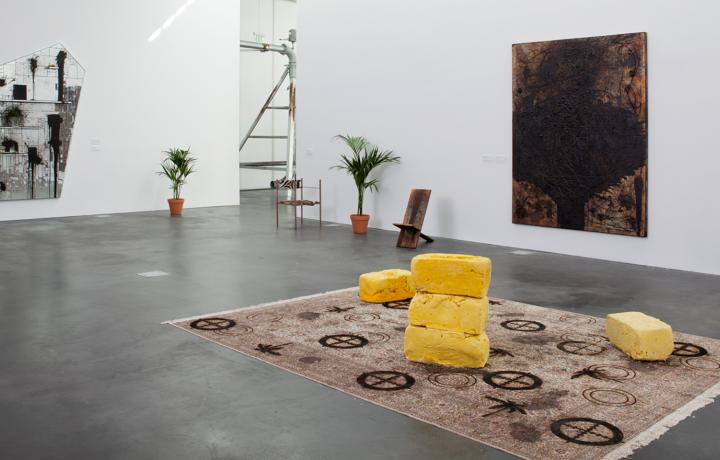 Installation shot in a gallery space with a concrete floor. Two large abstract paintings hang on the walls. In the center is a dirtied rug with large yellow bricks resting on top.  Two potted plants rest near the entrance of the gallery and two chairs are visible. 