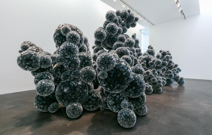 Installation shot of a Tara Donovan artwork in a gallery space. It is a large amorphous black sculpture composed of several spheres that catch the light resulting in an alien-like structure.