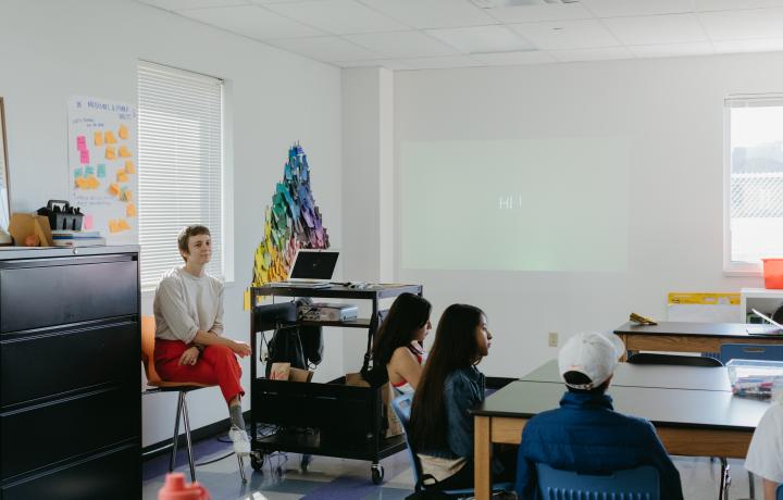 Image of artist Shannon Finnegan sitting at the front of a classroom, wearing red pants next to a colorful post it wall. And students sit at their desk watching the presentation