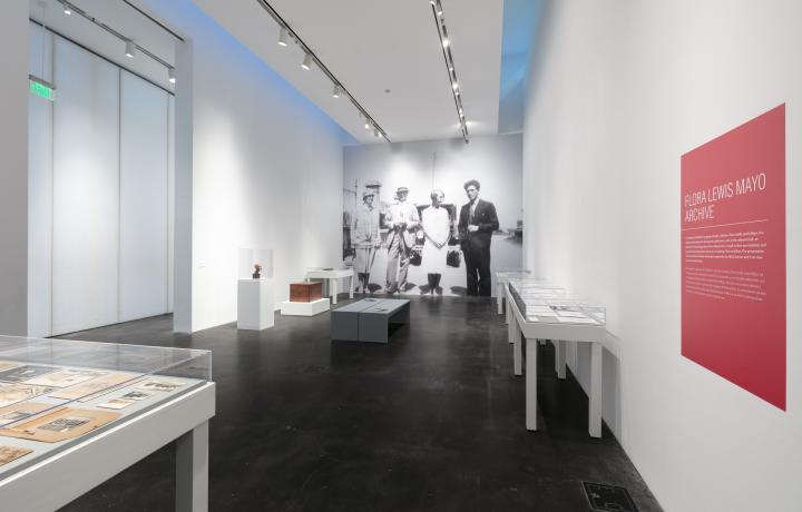Image of museum gallery. Far wall is covered in a black and white vinyl photograph depicting two men and two women dressed in attire from a different time. In the gallery are several tables displaying archival materials. 