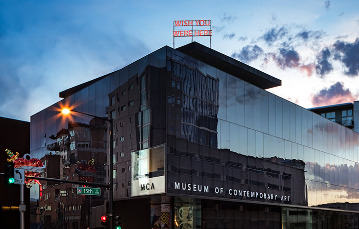 Image: Exterior shot of the Museum of Contemporary Art Denver at sundown. A neon sign in red is displayed on the buildings roof and reads "WISH YOU WERE HERE." The sunset is reflected on the buildings side.