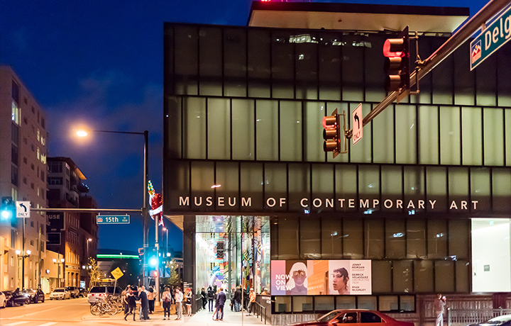 Image: Exterior shot of the Museum of Contemporary Art Denver at dusk. Several people gather near the entrance. Light glows through the glass building from within. ﻿