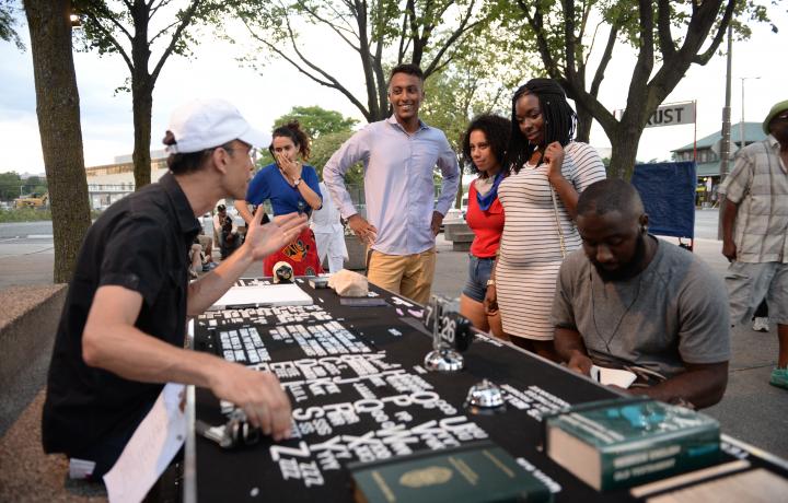 Candid shot of Paul Ramirez Jonas’ Public Trust activation. Paul is seated outdoors at a black table covered with letter tiles from A to Z, with multiple copies of each letter. He is surrounded by a ethnically diverse group of young adults which he is engaging with. 