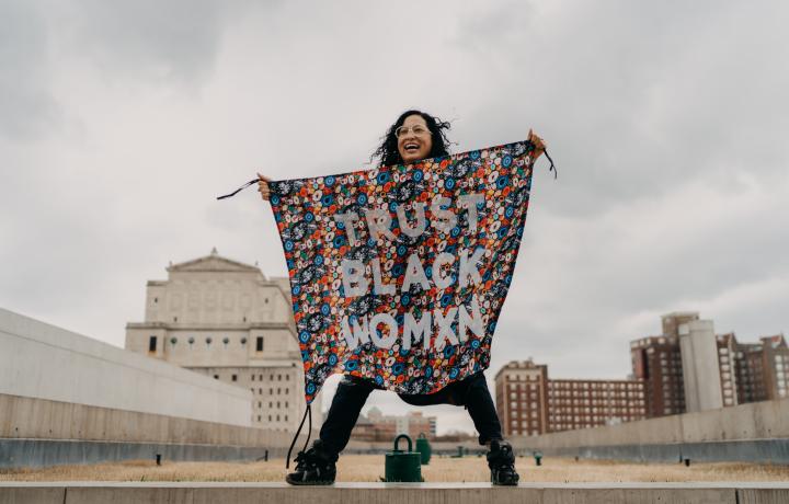  A photo of Aram Han Sifuentes, beaming. She’s holding a square, colorful banner that reads, “TRUST BLACK WOMXN”  in all capital letters. It appears as though she’s standing on the rooftop of a building, and a cloudy sky and tall buildings are behind her.