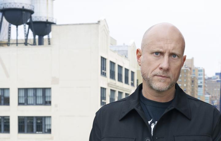 Portrait of Trevor Paglin against an urban landscape. He is wearing a dark coat as his cobalt eyes look sternly into the camera. 