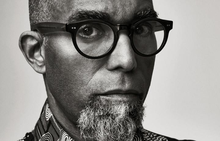 A black and white portrait of Dread Scott from the shoulders up against a neutral gray background. He looks seriously into the camera through his black rimmed glasses as a circular-patterned collar frames his salt-and-peppered beard and a tight black curly mohawk defines his upper face.