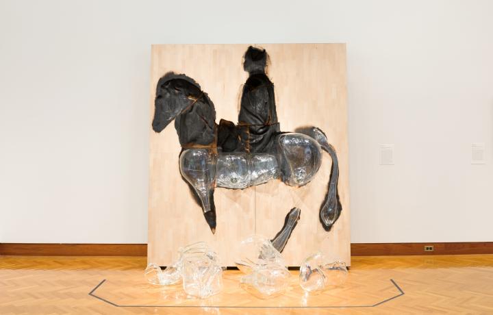  A large flat piece of wood rests against a wall in a gallery space. A figure of a man riding a horse is charred into the wood. Glass rests in the lower half of the charred spaces while on the floor of the gallery remains the upper portion of the horse and man.