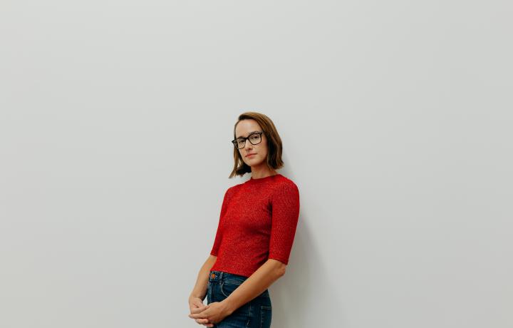 Portrait of Laura Shill leaning against a white background with her hair let down in a short brown bob. She is wearing a half-sleeve sweater that is the color of a red candy apple and blue jeans. Her hands are crossed in front of her as she looks softly into the camera.