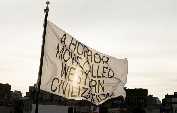 A white flag with the text “A HORROR MOVIE CALLED WESTERN CIVILIZATION” in all capital letters flies against an urban landscape. The sun is shining through the material of the flag.