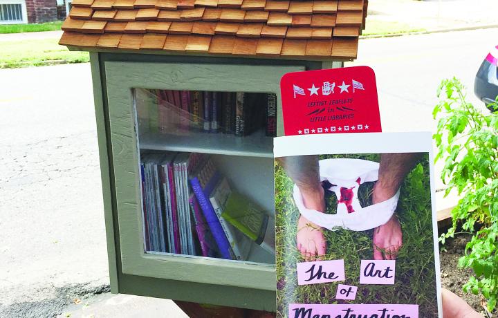 A hand is holding a leaflet reading “The Art of Menstruation” in front of a “little library” outdoors in a neighborhood. The image of the leaflet shows legs standing in grass with white underwear around their ankles; the underwear contains menstrual blood.