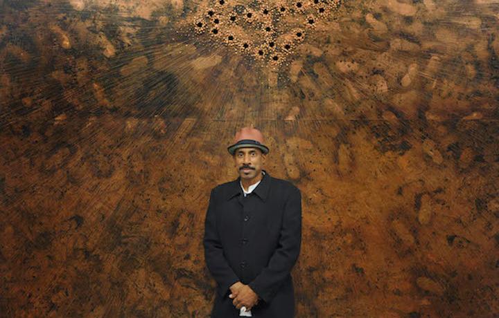 A portrait of Nari Ward taken from a slight distance against a large flat copper artwork of his. The artwork takes up the entirety of the frame with drilled holes making a diamond in the center. He stands proudly with his hands crossed while wearing a black jacket, a brown fedora.