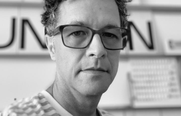 Black and white portrait of Paul, who is wearing glasses and a striped shirt. He has a serious look on his face, his hair is curly, and he is standing in front of some large text. 