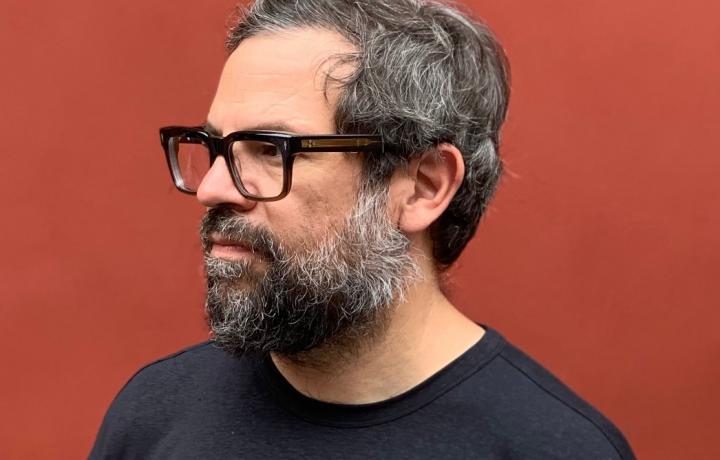 Portrait of Pedro Reyes staring off camera at a 45 degree angle standing against a solid rust-red backdrop. He is wearing rectangle rimmed glasses, a black t-shirt, and a silvering beard that connects to his hair. 