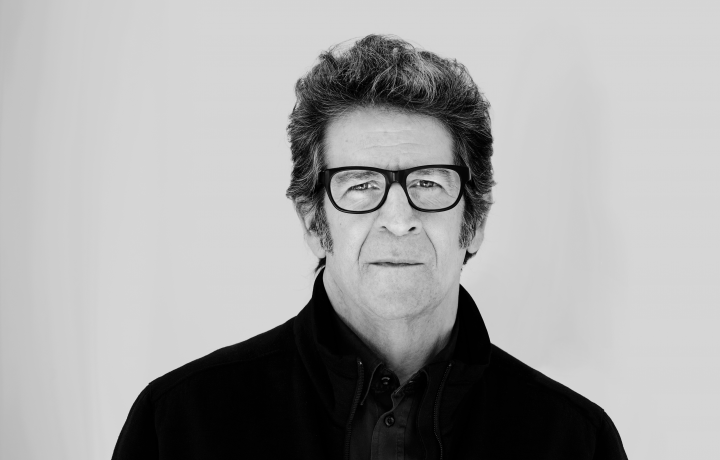 Black and white portrait of Robert standing against a plain wall. He has a serious look on his face, is wearing glasses and  a jacket over a button up shirt. 