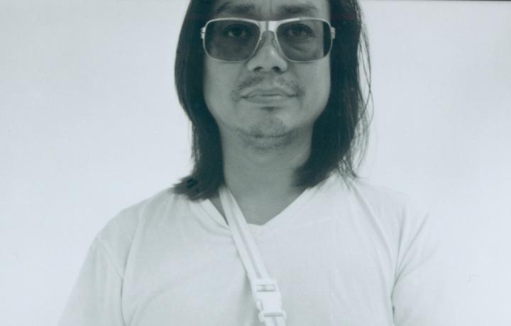 Portrait of Rirkrit Tiravanija against an off-white background in which his shirt seems to disappear into. His obsidian black hair is relaxed and meets his shoulder while metal framed sunglasses rest on his nose. He is looking just above the camera, calmly, with a neutral expression. 